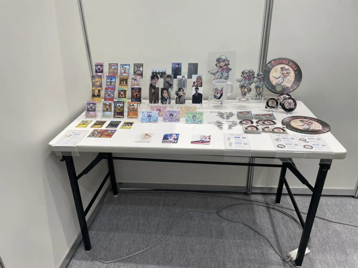 サンプル展示台に並べたARカード、アクリルスタンド、缶バッジ、マグカップ、レコード型ノベルティ